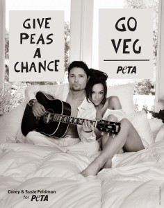 Corey Feldman and Susie Feldman in bed. Corey is playing guitar, sitting cross legged on the bed looking straight at the camera. His wife is leaning on him exposing much more skin.