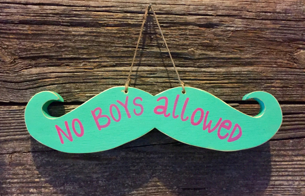 Hanging wall sign the shape of a mustache that reads, "No Boys Allowed"