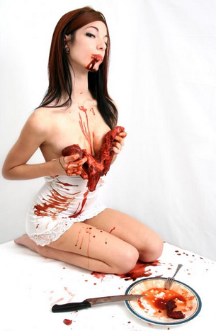 Thin white woman on her knees, kneeling in front of a plate with silverware and blood. She is rubbing bloody steaks over her breasts and looking seductively at the camera