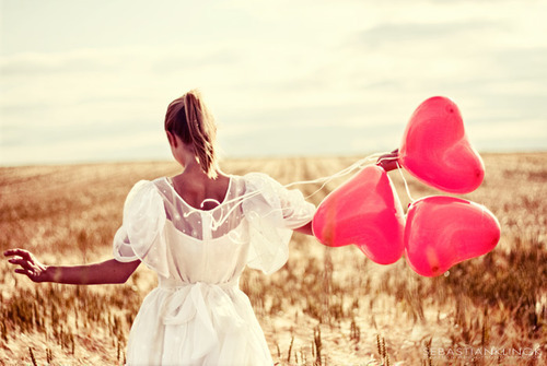 Woman in golden field and white dress holding heart shaped pink balloons