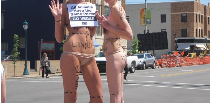 Two young thin white PETA volunteers pose naked on a street corner with their bodies marked like meat cuts holding a PETA sign that asks viewers to go vegan