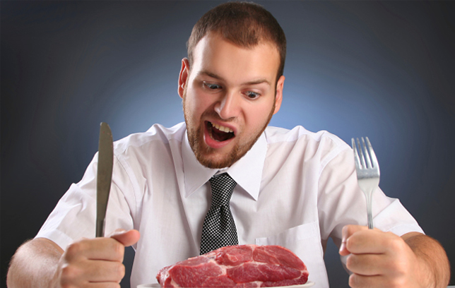 Man in a suit sits in front of a plate with a raw steak, knife and fork poised in his fists on the table